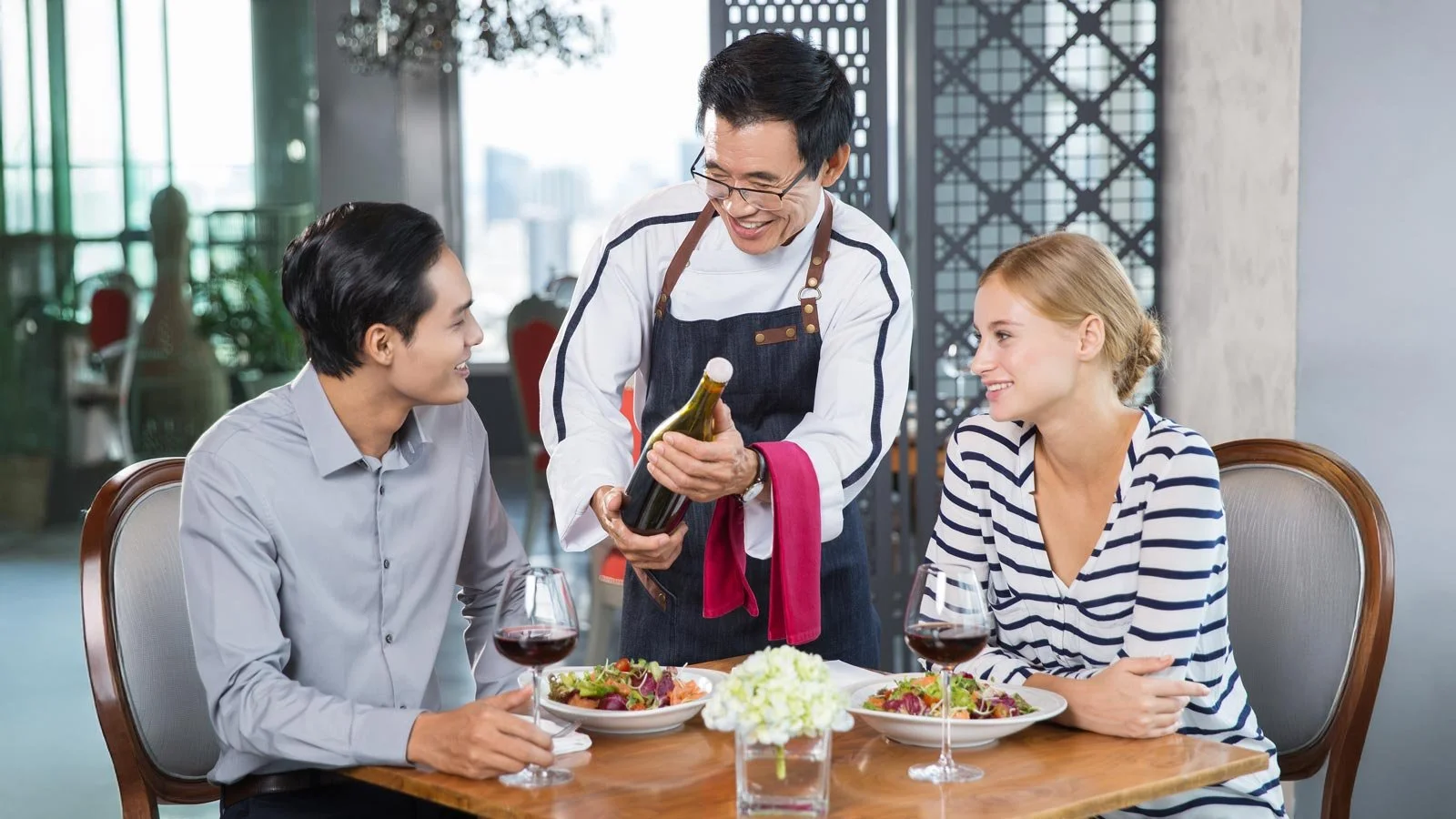 Smiling Asian Waiter Offering Wine to Young Couple